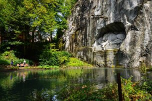 A rock face with a lion sculpture carved into it overlooks a pond, surrounded by lush greenery. Two people stand on the path beside the pond, enjoying one of the top things to do in Lucerne amidst this serene setting.
