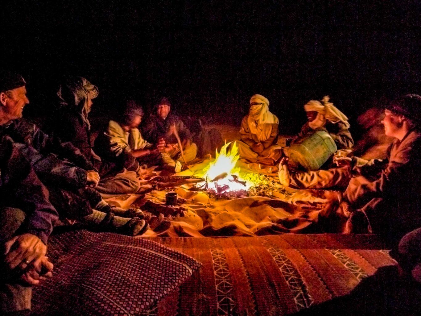 Nighttime in the Moroccan desert with a group of robed and turbaned nomads gathered around a roaring campfire.