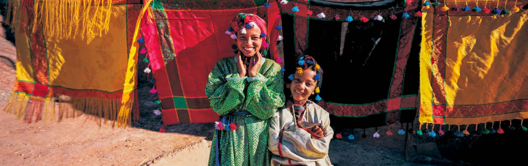 Two Moroccan girls with big smiles and colorful clothing stand in front of bright textiles.
