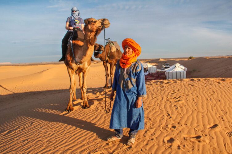 Cameleer in blue robe and bright orange turban leads guest on a camel with luxury Sahara Desert camp in the background.