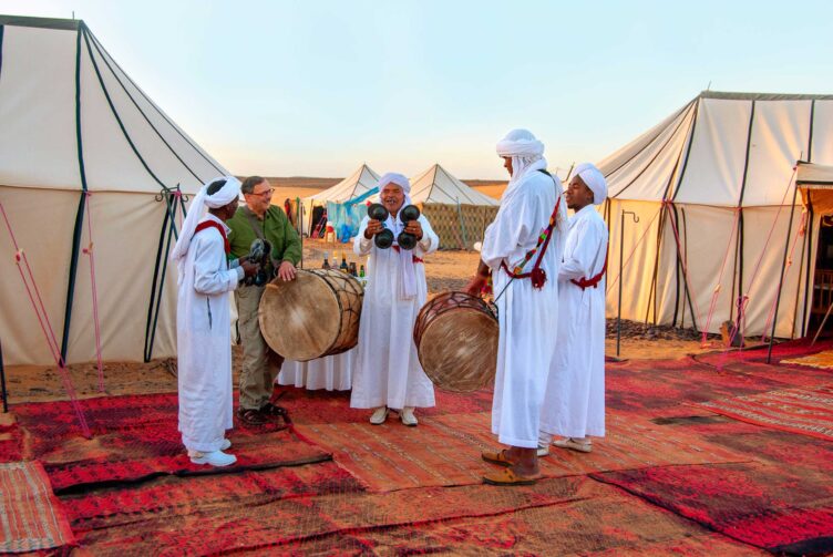 Four white-robed musicians talk with traveler in front of large white tents at luxury Sahara Desert camp.