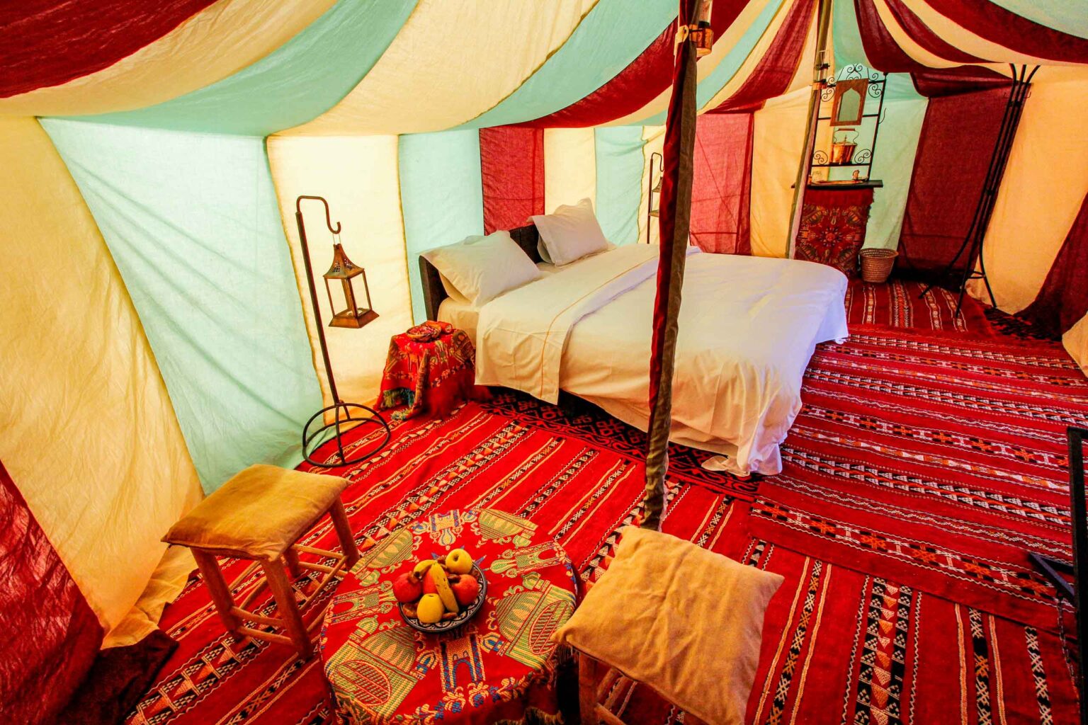 Well lighted walk-in tent with double bed and a floor covered with red Moroccan carpets.
