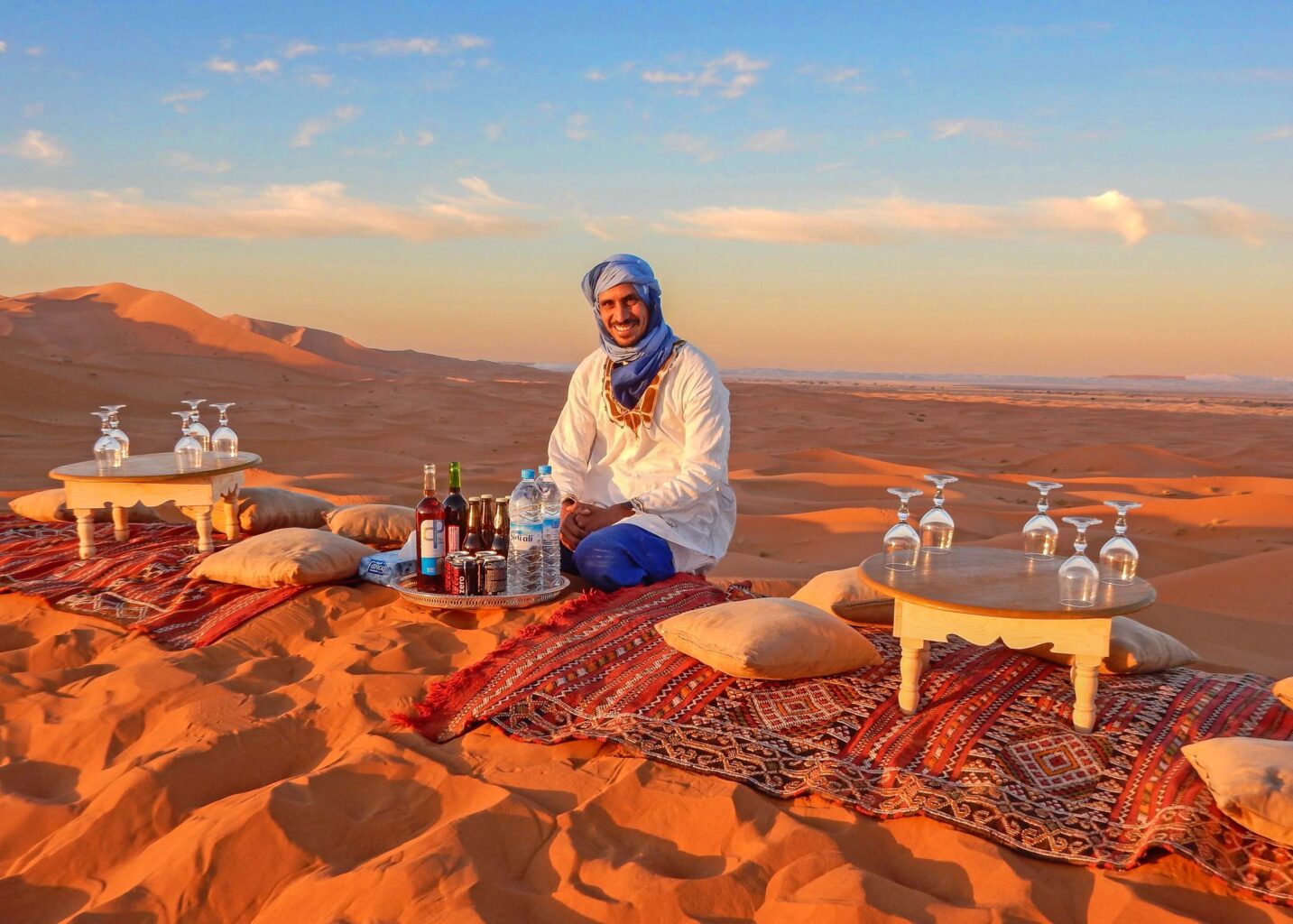 Smiling host in a white robe and blue turban offers refreshing beverages with Sahara Desert dunes in the background.