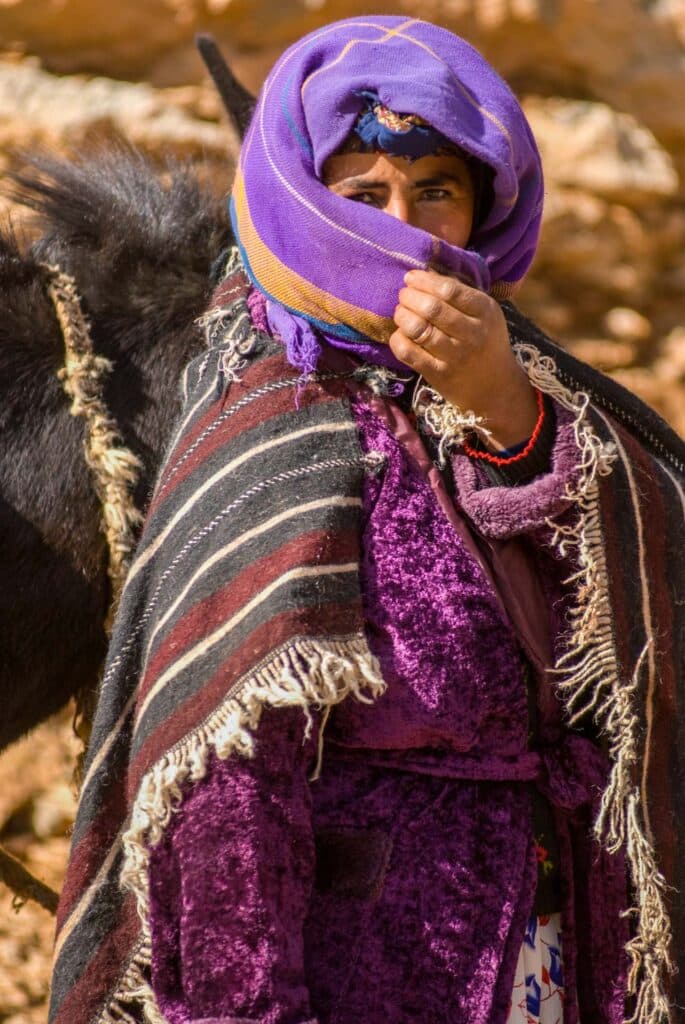 Moroccan nomadic woman in purple clothing looks out from behind her face-covering hijab as she leads a black horse.