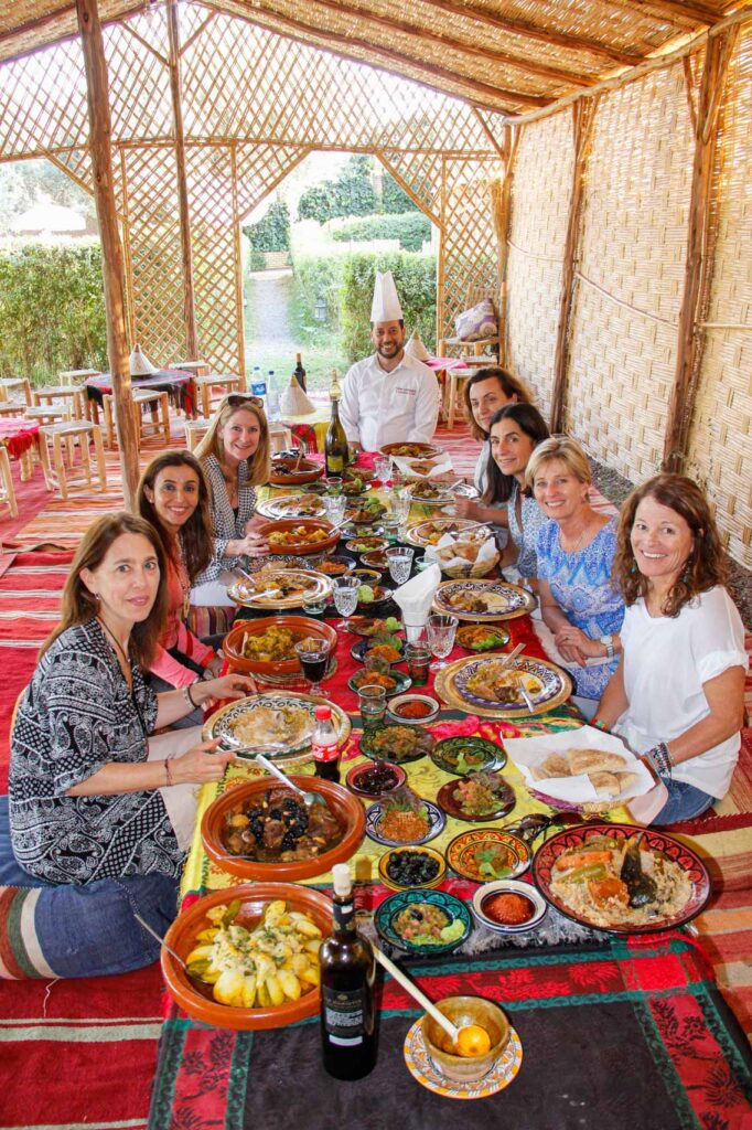 Eight travelers and a chef smile at a table overflowing with many plates of colorful, delicious looking Moroccan cuisine.