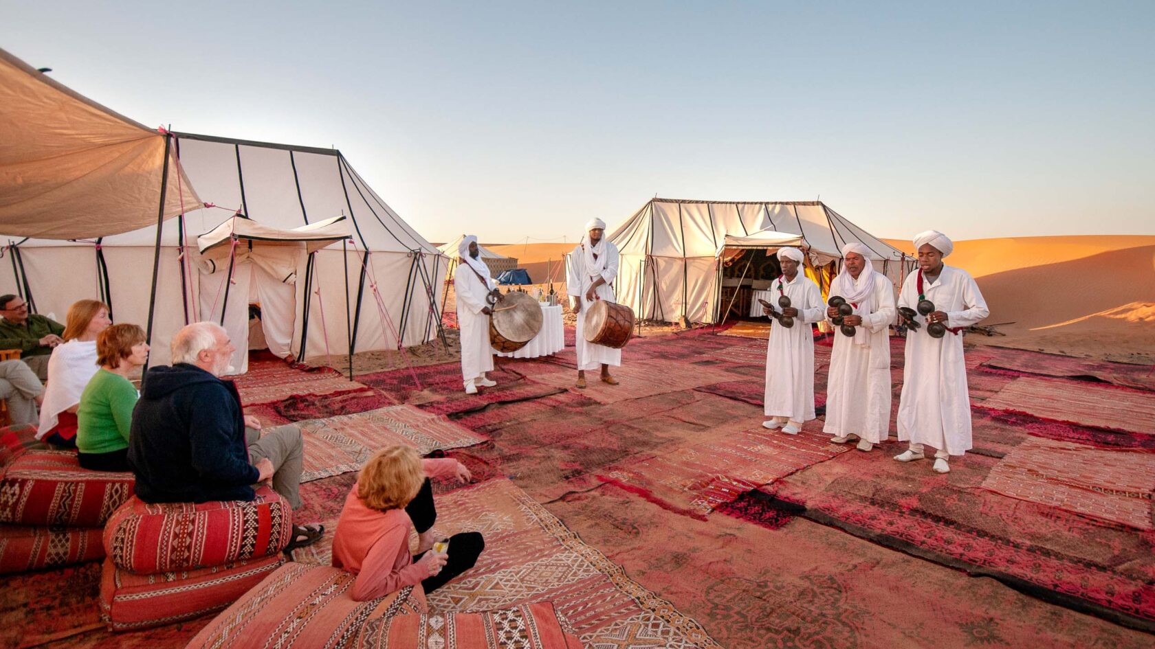 Travelers lounge on Moroccan trugs and futons as they listen to white-robed musicians in the Sahara Desert.