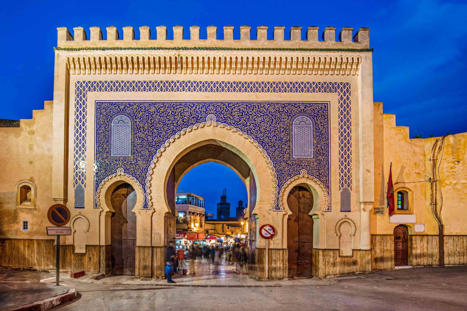 Grand crenellated entrance gate to a Moroccan city is covered in blue geometric tiles in front of bright blue sky.