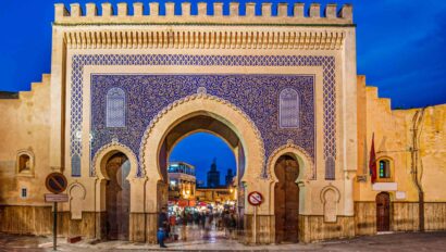Grand crenellated entrance gate to a Moroccan city is covered in blue geometric tiles in front of bright blue sky.