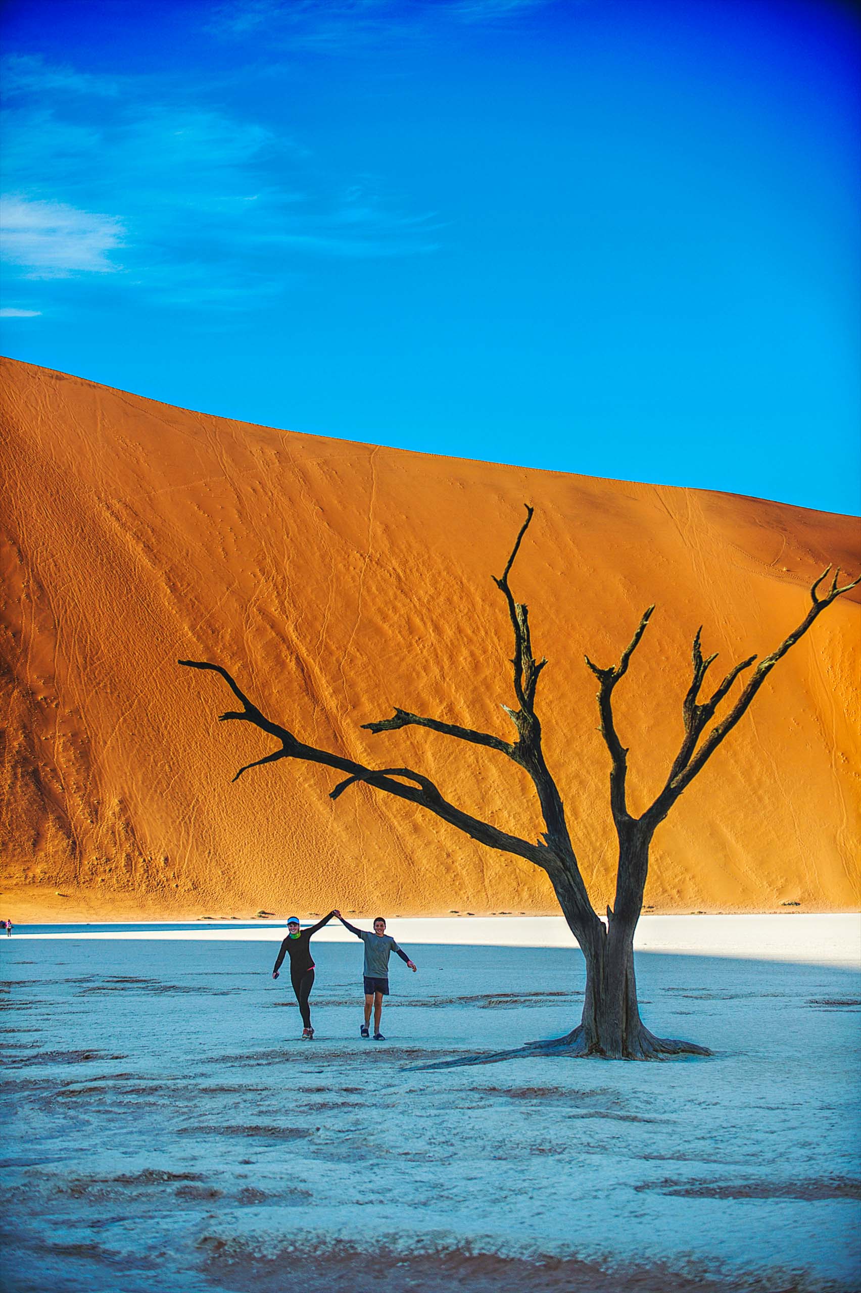 A young girl and young boy holding hands running together over the clay salt pan smiling between the Dead Vlei trees.