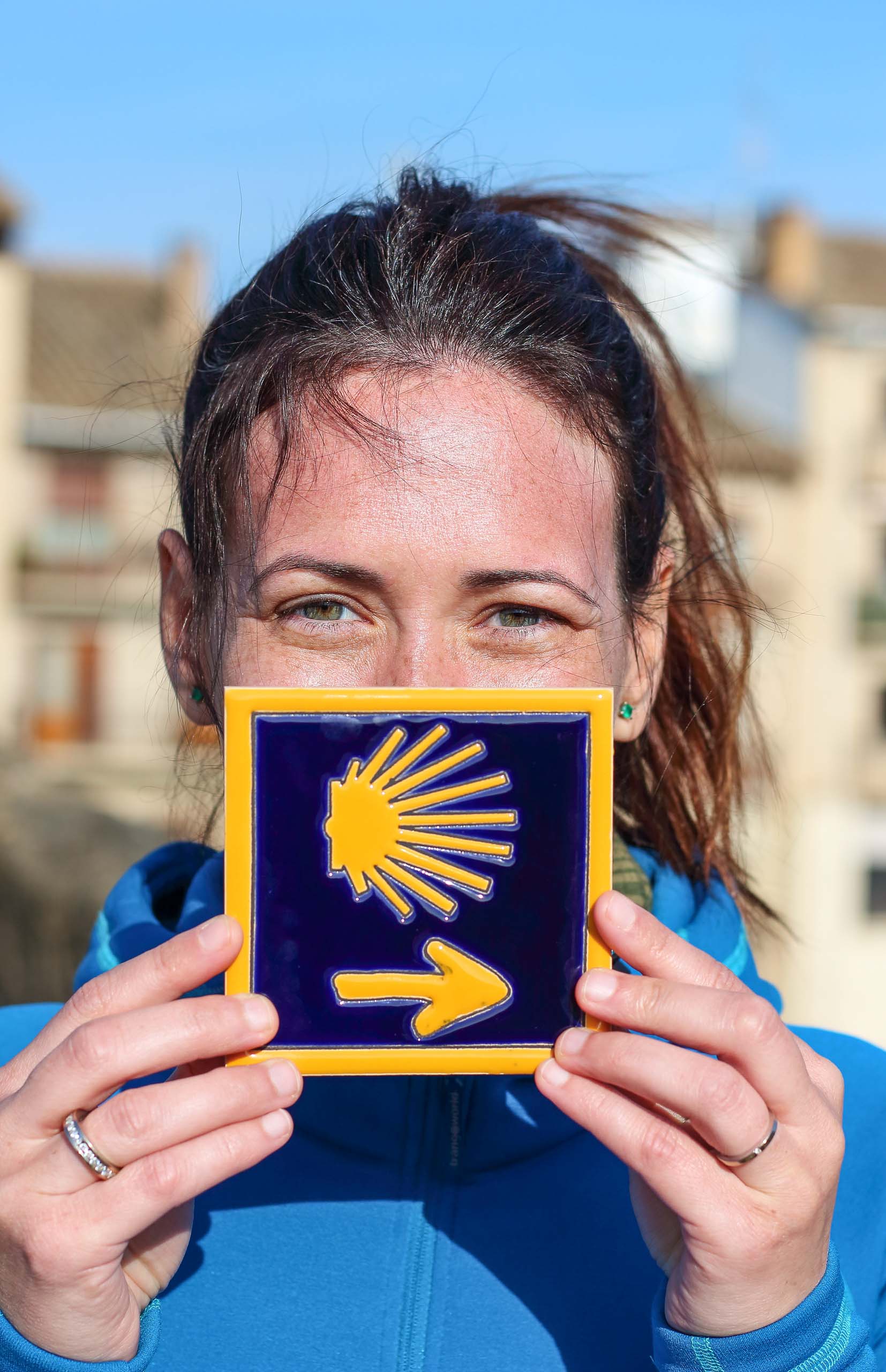 A woman holding a "Camino de Santiago" blue tile painted with a yellow shell and arrow.