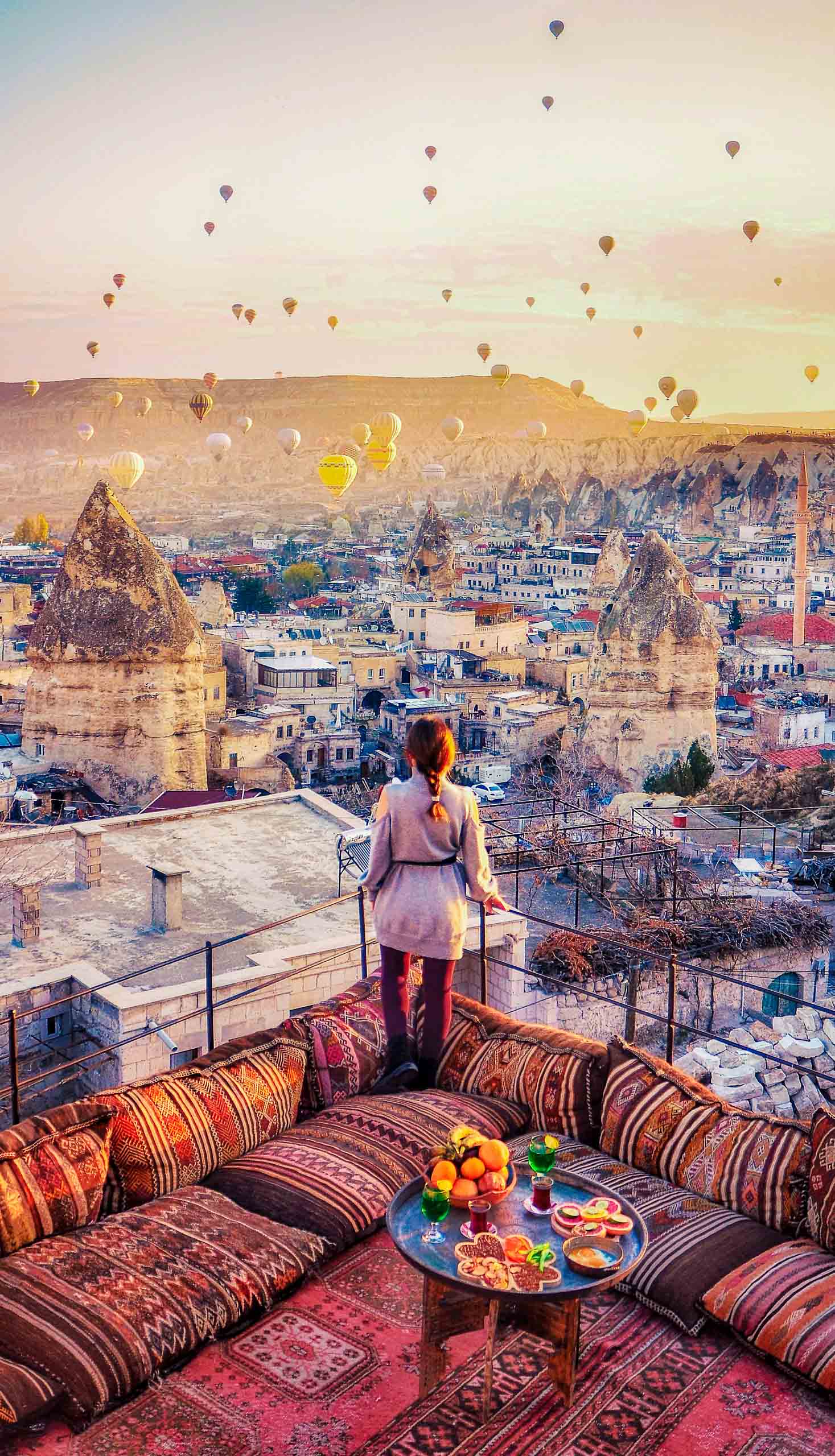 A woman stands at hotel rooftop watching hot air balloons flying over a city in Turkey.