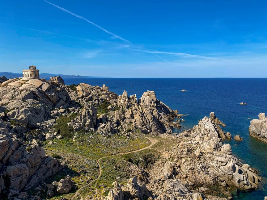 A rocky coastal landscape with a small circular building on a hill overlooking the sea under a clear blue sky, reminiscent of the adventures one might find in Corsica or Sardinia.