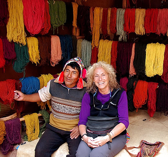 Two people sitting in front of shelves filled with various colored yarns. One person wears a traditional hat and the other is smiling, surrounded by the cozy threads spun from alpacas.