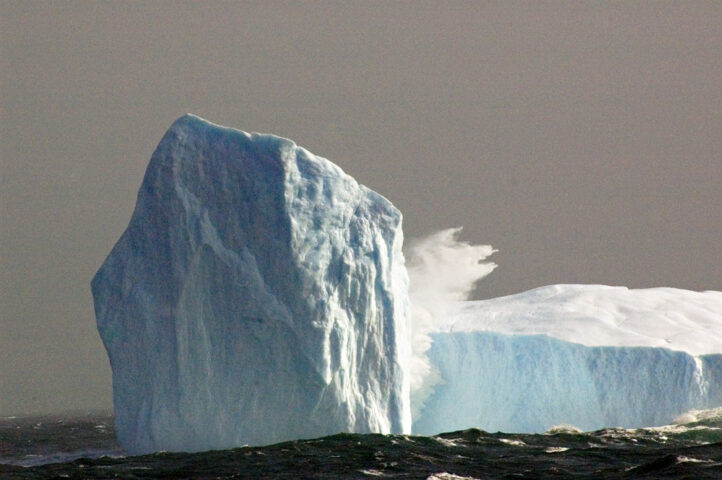 A large iceberg with a steep, jagged peak rises from the ocean. Waves crash against its base under a cloudy sky, a true blue icebreaker in this icy wilderness.