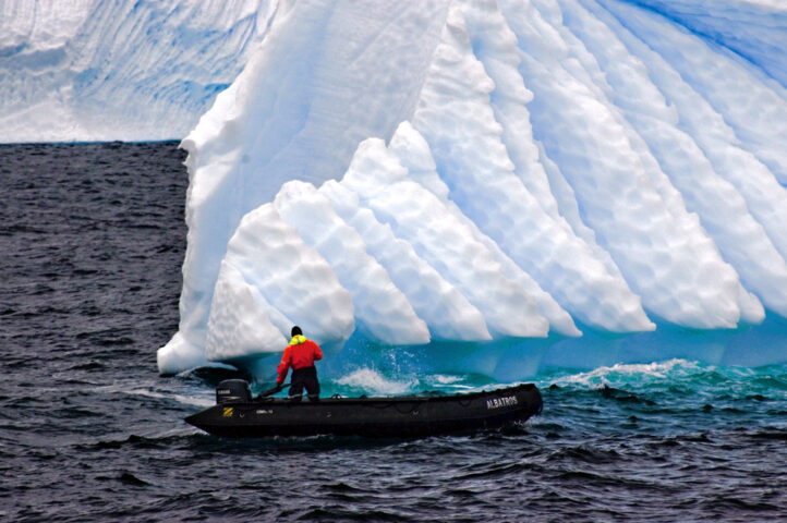 A person in a small motorboat navigates close to a large, snow-capped iceberg in dark waters, their True Blue jacket standing out against the icy backdrop.