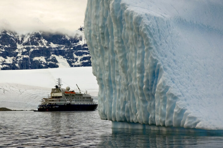 A true blue icebreaker rests in the foreground, while a small cruise ship lingers in the distance. Snow-covered mountains rise majestically under a cloudy sky.
