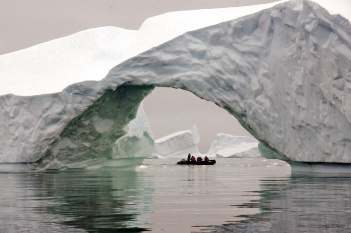 A small boat with people, aptly named "True Blue," passes through an arch in a large iceberg on calm waters, resembling an icebreaker navigating serene polar landscapes.