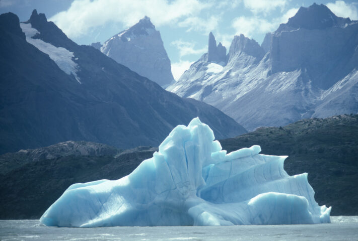 A large iceberg floats in the foreground with jagged, snow-capped mountain peaks in the background under a partly cloudy sky, showcasing nature's true blue palette.