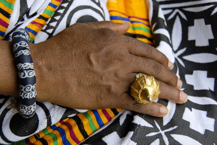A hand, evoking the grandeur of royalty, adorned with a large gold ring and a black and white patterned bracelet, rests on multicolored, patterned fabric.