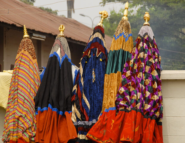 Colorful, elaborately designed traditional costumes with tall, cone-shaped hats are displayed outside, leaning against a white wall. Trees and a corrugated metal roof in the background subtly hint at the rich Ashanti culture.