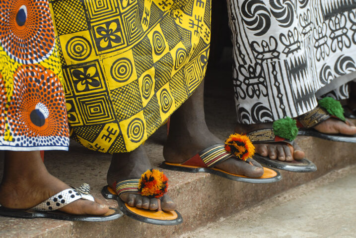 Four individuals, reminiscent of the Ashanti King and I, are wearing colorful, patterned fabrics and traditional sandals adorned with decorations as they stand on a stone surface.