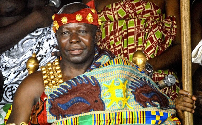 A man dressed in traditional, colorful Ghanaian clothing and jewelry, wearing a headdress reminiscent of an Ashanti King, sits among a group of people.