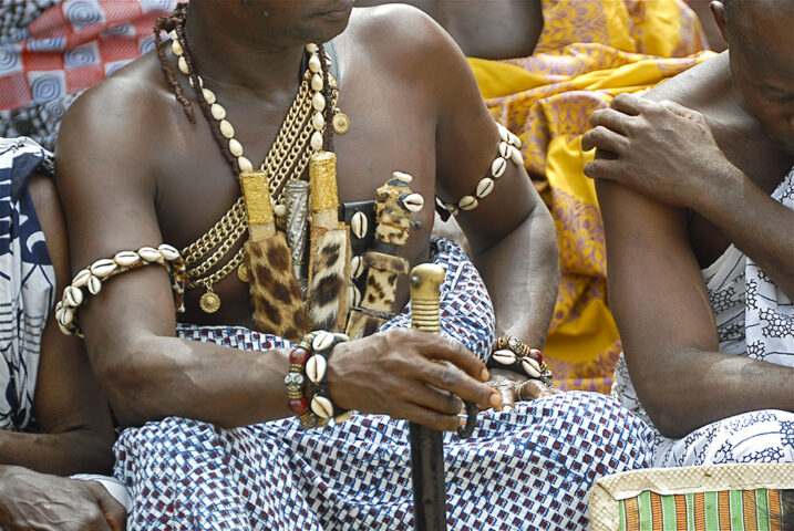 A person dressed in traditional attire adorned with beads, shells, and leopard skin patterns holds a staff, reminiscent of an Ashanti King. Other attendees in similar clothing are partially visible around them.