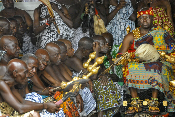 A group of traditionally dressed men surrounds the Ashanti King, who is seated on an ornate throne. The men are in colorful patterned attire, with some holding ceremonial objects, creating a vibrant scene reminiscent of "Ashanti King and I.