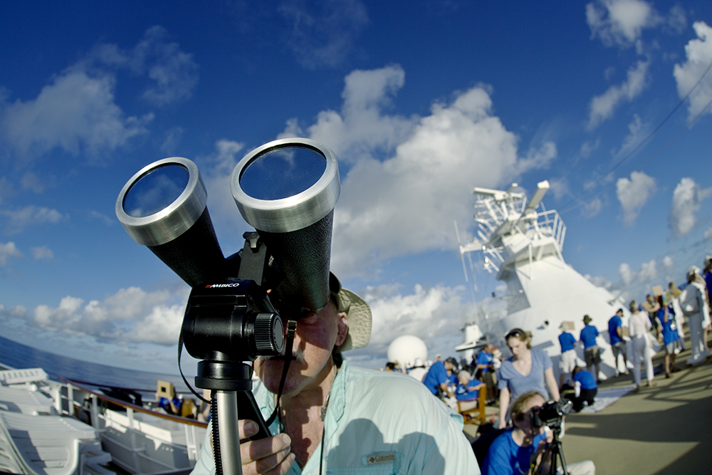 A person on a ship looks through large binoculars, marveling at the amazing sight in the universe, with other people and nautical equipment visible in the background under a blue sky with clouds.