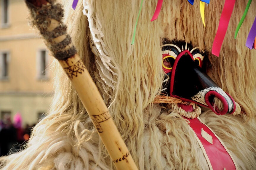 A person in a traditional Slovenian-style costume with long fur, a colorful mask, and feathers, holds a decorated wooden stick.