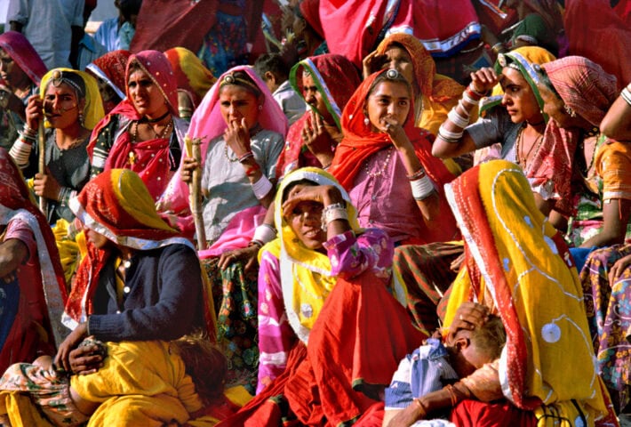 A group of women dressed in vibrant traditional clothing and headscarves sit together outdoors, some shading their eyes with their hands, evoking the atmosphere of an exotic place perfect for world travel adventures.