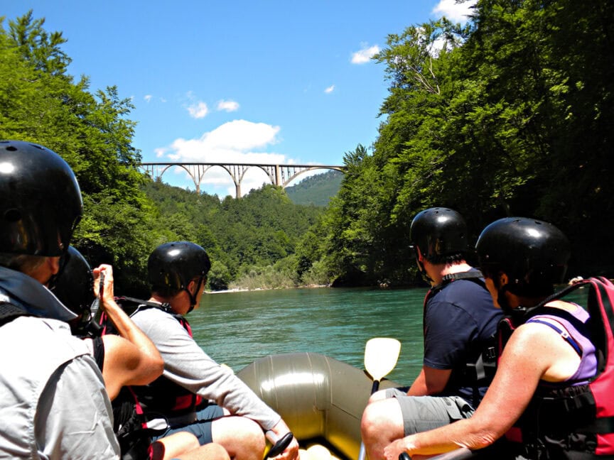 A group of people wearing helmets and life jackets are rafting on a river. Ahead, there is a large bridge framed by lush green trees and a clear blue sky, leading them into the adventure of the Tiger's Nest.