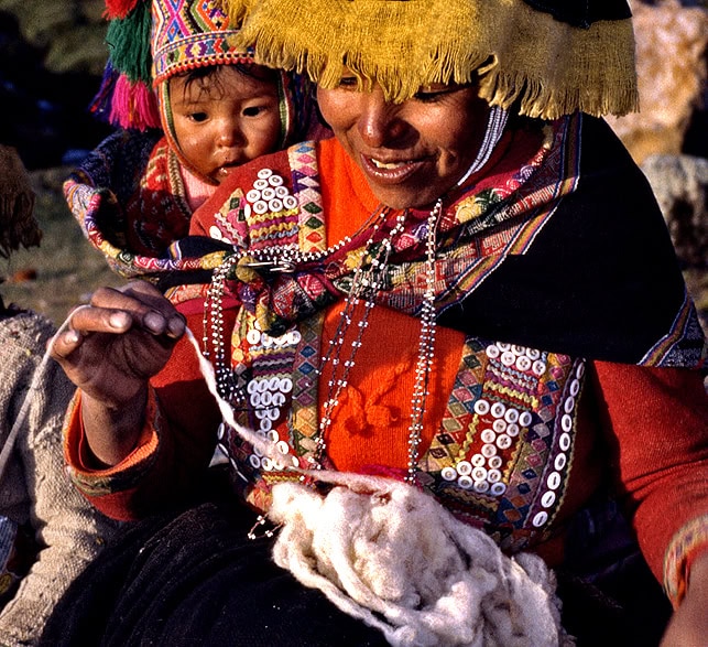 A person in traditional colorful attire spins wool with a spindle while carrying a child on their back, as llamas graze peacefully in the background.