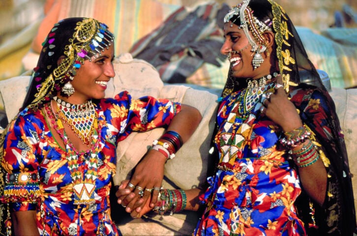 Two women dressed in colorful traditional attire and elaborate jewelry smile at each other while holding hands outdoors, capturing the essence of an exotic destination.