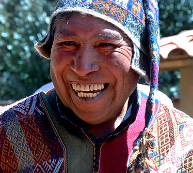 A man wearing a colorful knit hat and traditional patterned clothing smiles widely, showing his teeth. A blurred natural background with hints of Alpacas is visible behind him.