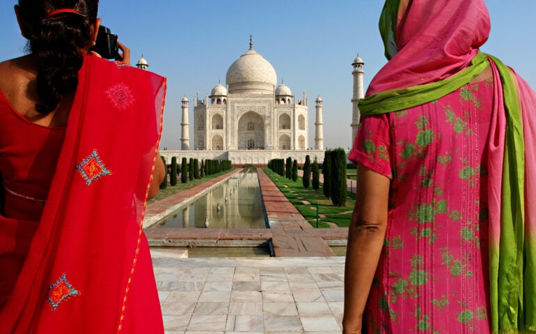 Two people in colorful traditional attire stand facing the Taj Mahal, one taking a photo. The white marble mausoleum, considered one of the most exotic places in the world, is centered in the background, with clear skies and greenery surrounding it.