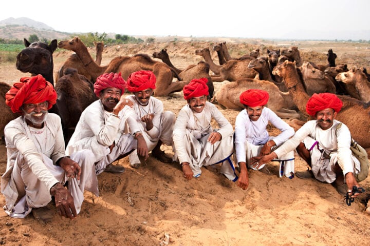 Five men wearing white clothing and red turbans sit on the sand with camels resting in the background, capturing the essence of an exotic place that's a perfect travel destination.
