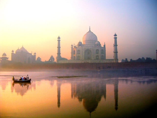 A small boat with passengers floats on a river at sunrise, with the Taj Mahal in the background reflecting in the water—truly an exotic destination for world travel.