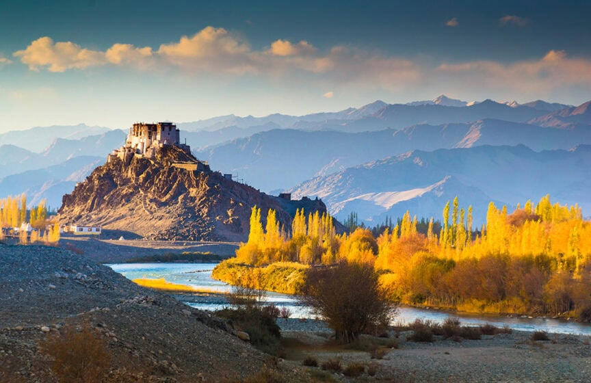 A monastery sits atop a rocky hill overlooking a river and colorful autumn foliage, with mountain ranges visible in the background under a partially cloudy sky. This hidden gem was among the top 10 travel destinations of 2017, offering breathtaking views and serene beauty.