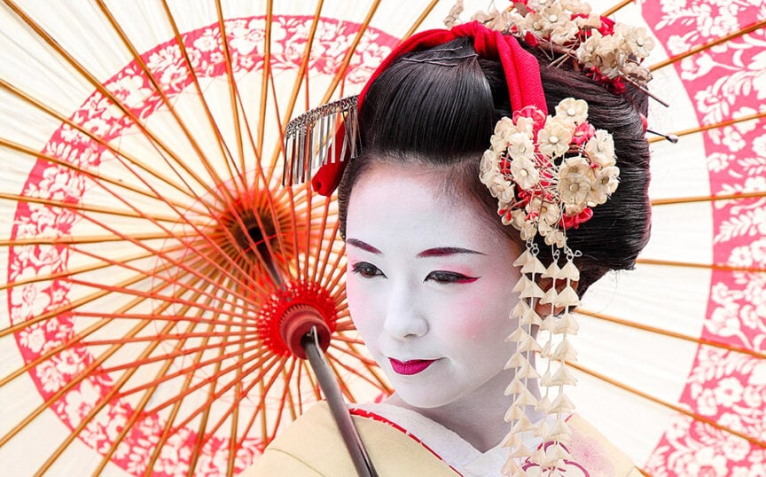 A woman dressed as a traditional Japanese geisha holds an ornate umbrella. She wears white face makeup, red lipstick, and floral hair accessories, embodying the charm of one of the best places to visit in 2017.