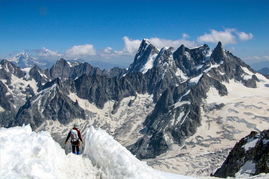 A mountaineer stands on a snow-covered peak, overlooking a rugged mountain range with sharp, jagged peaks under a clear blue sky. It's as if they are charting the Haute Route from France to Switzerland, ready for an epic hiking adventure.