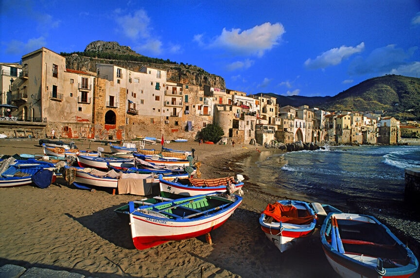 A coastal village with colorful boats on a sandy beach, stone buildings in the background, and a rocky hillside covered in greenery under a blue sky with scattered clouds. This picturesque destination is among the top 10 places to travel for those seeking serene beauty and charm.