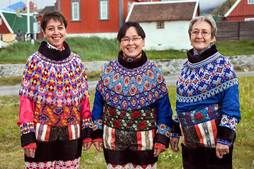 Three people are standing outdoors, wearing colorful traditional clothing with intricate patterns. They are smiling with red wooden buildings in the background—truly capturing the essence of one of 2017's top travel destinations. If you're curating your list of places to travel, this should be on it!