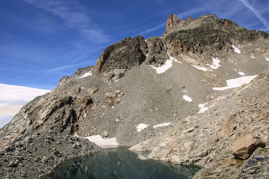 Rocky mountain landscape with patches of snow and a small lake, under a clear blue sky—perfect for hiking along the scenic Haute Route from France to Switzerland.
