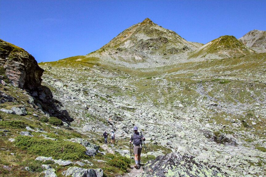 Hikers walk along the rugged Haute Route trail towards a steep, green and brown mountain peak under a clear blue sky.
