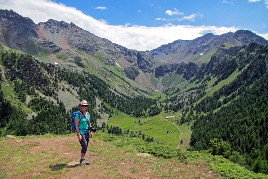 A person in hiking attire stands on a trail, overlooking the lush green Val d'Aosta valley in Italy, surrounded by rugged, tree-covered mountains.