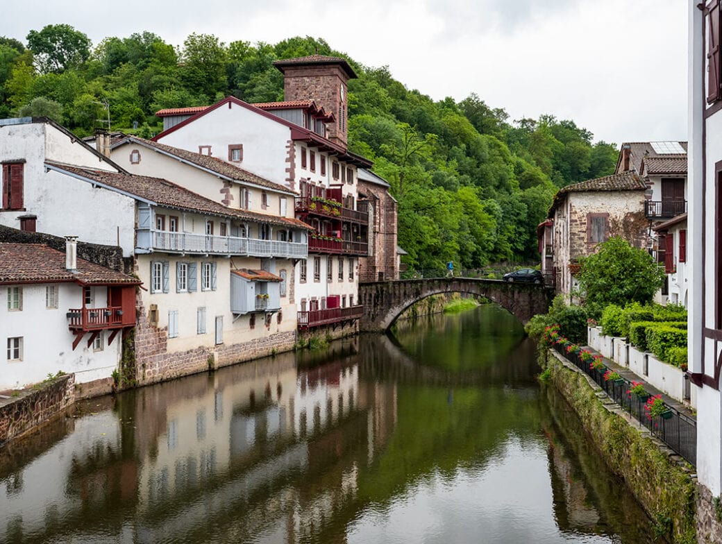 A narrow river flows through a quaint village with white and brick buildings and a stone bridge, surrounded by lush greenery, perfect for hiking enthusiasts exploring the serene Basque Country.