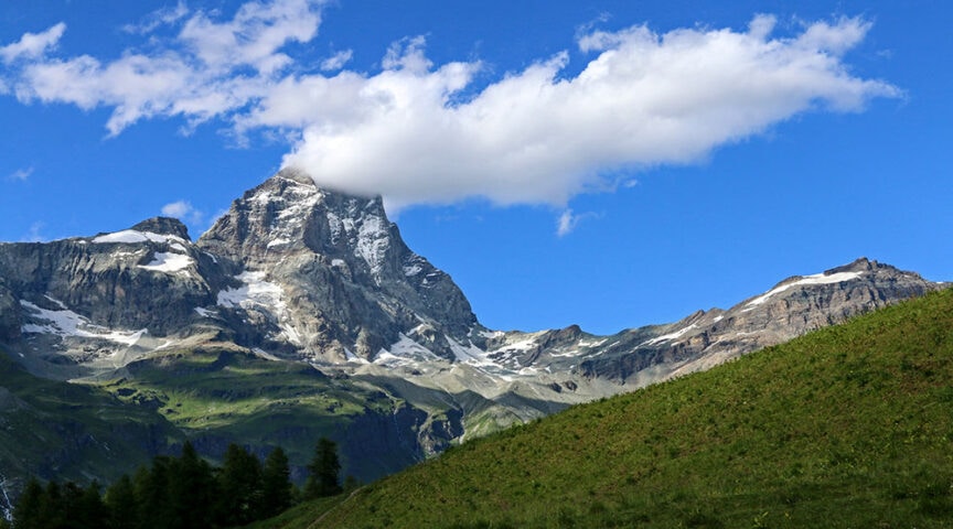 A mountain peak in Val d'Aosta, Italy with a cloudy sky above, partially covered by a single cloud, surrounded by green slopes and patches of snow—perfect for hiking enthusiasts.