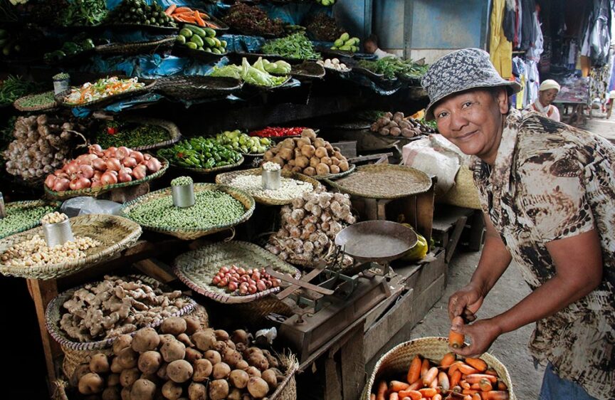 A vendor wearing a hat and patterned shirt smiles while arranging produce at a market stall filled with a variety of vegetables, evoking the feeling of an island lost in time.