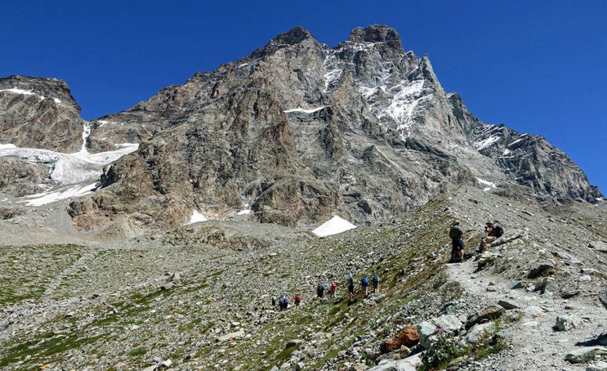 A group of hikers walk along a rocky trail at the base of a tall, rugged mountain under a clear blue sky in the stunning Val d'Aosta, Italy.